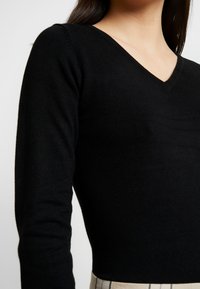 Černý pletený top s dlouhým rukávem a výstřihem do V, se přiléhavým střihem a hladkou texturou. Žádné viditelné vzory ani akcenty.
