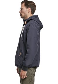 Chaqueta con capucha de color azul marino, con cremallera en la parte frontal, cordones blancos, puños elásticos y un ajuste relajado. Se aprecia un bolsillo lateral con cremallera.