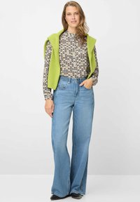 Gilet verde lavorato a maglia sopra una camicetta stampata con collo alto, abbinati a jeans a gamba larga di colore azzurro chiaro. Scarpe nere completano l'outfit.