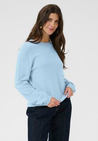 Hosszú barna hajú nő, aki arany fülbevalót visel, világoskék hosszúujjú pulóvert és sötétkék nadrágot, mindkét kezével a pulóver alját fogja.