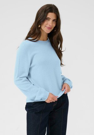 Frau mit langen braunen Haaren, die goldene Ohringe trägt, einen hellblauen langärmligen Pullover und dunkle blaue Hosen anhat, während sie den Saum ihres Pullovers mit beiden Händen hält.