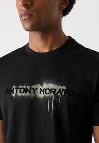 Tricou negru din bumbac cu numele brandului "ANTONY MORATO" în stil spray paint, având un accent texturat, de culoare verde estompat. Mâneci scurte.