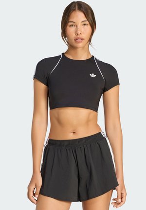 Jeune femme portant un haut de sport court noir et un short de sport noir avec des bandes blanches, debout devant un fond uni.