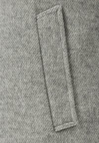 Grauer Wollstoff mit diagonaler Textur und einem rechteckigen Aufnäher auf der Oberfläche. Texturiertes und warmes Erscheinungsbild.