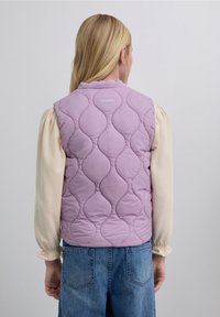 Veste matelassée violet clair avec motif en forme d'ondulations, marquée du nom "SCALPERS" au dos, portée avec une chemise crème à manches longues et un jean.