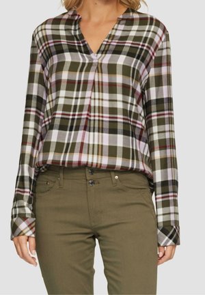 Woman wearing long-sleeve plaid shirt in green, white, and red tones paired with olive green high-waisted pants.