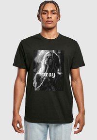 Sort t-shirt med et frontgrafik af en kvinde med langt hår og tatoveringer, der holder hænderne sammen med ordet "pray" nedenunder.
