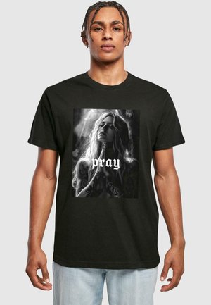 Sort t-shirt med et frontgrafik af en kvinde med langt hår og tatoveringer, der holder hænderne sammen med ordet "pray" nedenunder.