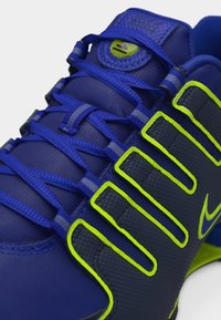 Κοντινή λήψη αθλητικού παπουτσιού Nike Shox σε μπλε και μαύρο χρώμα με νέον πράσινες λεπτομέρειες και αναλυτικά κορδόνια.