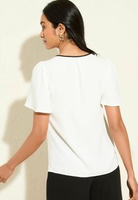 Blusa bianca con maniche corte e voluminose, bordino nero al colletto, texture liscia e una leggera forma ad A. Abbinarla a pantaloni neri.