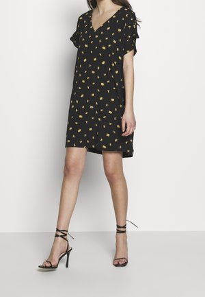 Robe noire à manches courtes et col en V, ornée d'un motif floral jaune. Assortie à des sandales à talons noires avec des lanières nouées à la cheville.