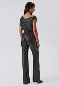 Jumpsuit nero con paillettes, dal collo drappeggiato, pantaloni a gamba larga e vita aderente. Presenta una texture scintillante e sottili pattern verticali.