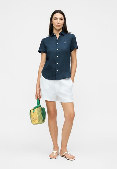 Γυναίκα που φοράει μπλε κοντομάνικο πουκάμισο με κουμπιά, λευκά σορτς, λευκά σανδάλια, κρατώντας μια πράσινη και κίτρινη πλεκτή τσάντα.