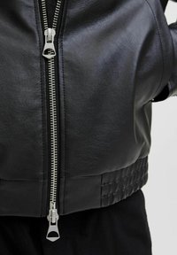 Chaqueta de cuero negro con un cierre central plateado, detalle acanalado en el dobladillo y textura suave. Primer plano de la tela y los herrajes.