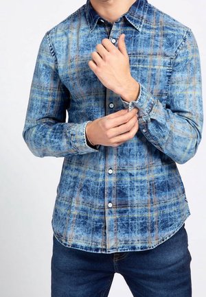 Mężczyzna zapinający mankiet niebieskiej koszuli w kratę z długim rękawem, w połączeniu z ciemnoniebieskimi dżinsami na jednolitym tle.