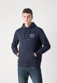 Navy blå hettegenser laget av myk stoff, med en frontlomme (kangaroo pocket), snor til hetten og trykt logo på brystet.