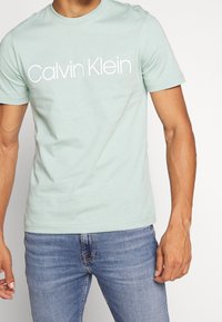 Camiseta de algodón verde claro con mangas cortas, que presenta un logo blanco de "Calvin Klein" en el pecho. Ajustada, con textura suave.