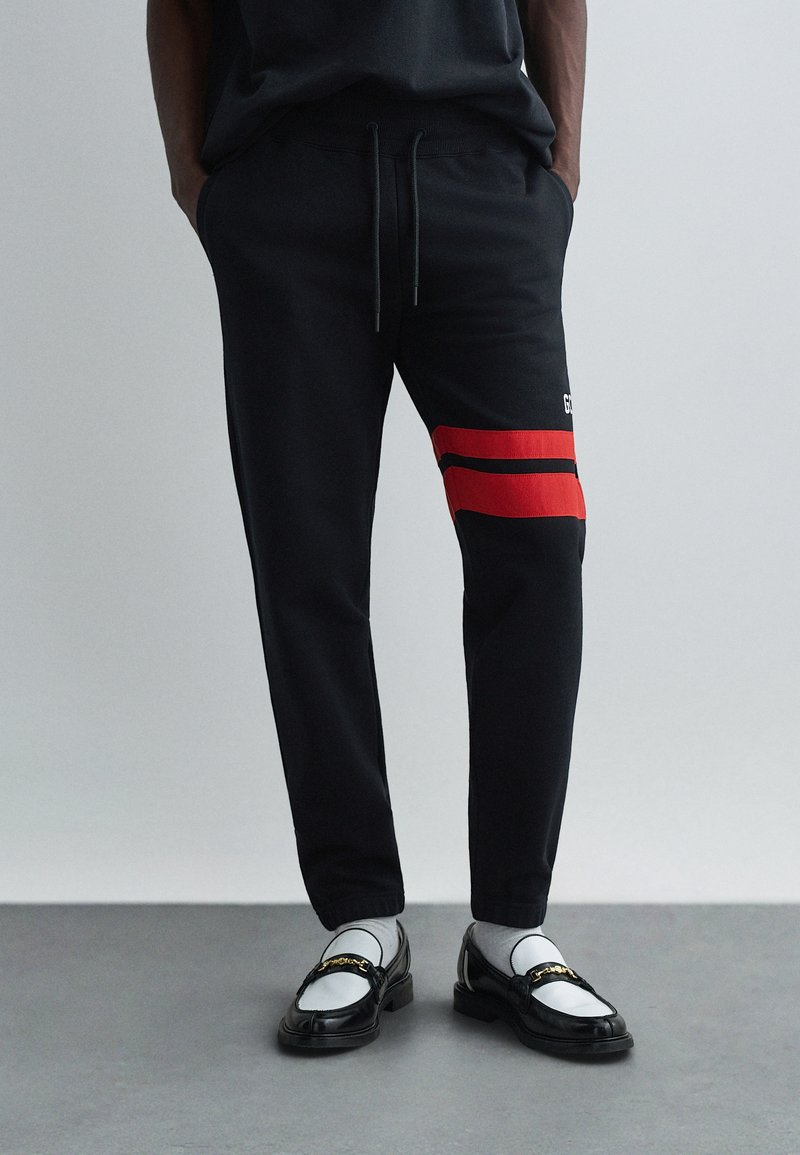 Svarta sweatpants med röda ränder, i bomull, med elastisk midja och dragsko, tillsammans med svarta loafers och grå strumpor.