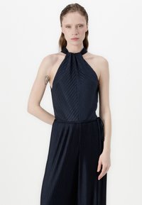 Navy halter jumpsuit with pleated texture, featuring a fitted bodice and flowing wide-leg pants, tied at the waist with a matching fabric belt.