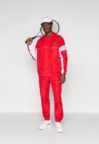 Κόκκινο αθλητικό κοστούμι με λευκές και μπλε λεπτομέρειες, ελαφρύ υλικό, με φερμουάρ και ελαστικούς καρπούς, φορεμένο με λευκά αθλητικά παπούτσια και καπέλο.