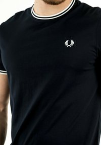 Camiseta de algodón de manga corta negra con un cuello acanalado que presenta un borde blanco y un pequeño logotipo de laurel bordado en el pecho.