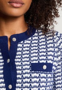 Granatowo-biały teksturowany kardigan z guzikiem przy dekolcie, wyposażony w kieszeń na piersi oraz ozdobne guziki.
