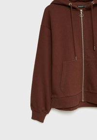 Bruine zip-up hoodie met een capuchon, oversized pasvorm, elastische manchet- en zoomafwerking. Heeft voorzakken en metallic ritsdetails. Zachte stoftextuur.