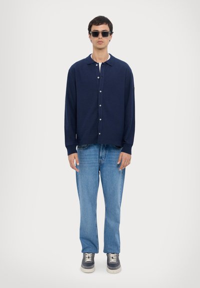Cardigan navy con bottoni e colletto, abbinato a jeans dritti azzurri chiari e sneakers scure. Modello con occhiali da sole.