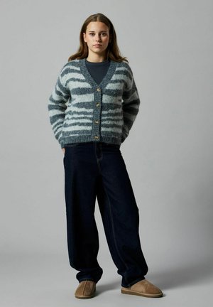 Cardigan en tricot à rayures grises et blanches, avec un col en V et cinq boutons dorés, associé à un jean large noir et des chaussures à enfiler marron clair.