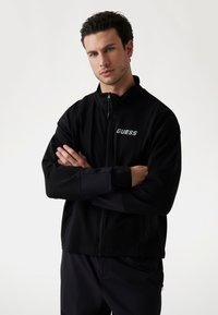 Svart zip-up jacka med GUESS-logotyp på bröstet, med hög krage och två sidofickor, tillverkad av mjukt tyg med en slät yta.