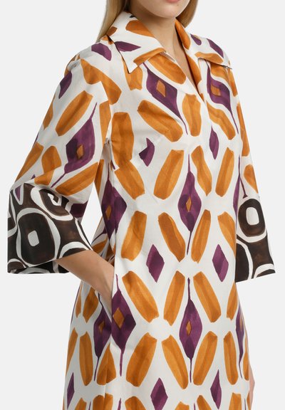 Abito con maniche in tessuto bianco, caratterizzato da una stampa geometrica arancione, viola e marrone. Il design include un colletto classico e tasche laterali.