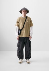 Beige bomulds T-shirt lagd over en hvid langærmet skjorte, sorte cargo-bukser med lommer, grå bredskygget hat og beige sneakers.
