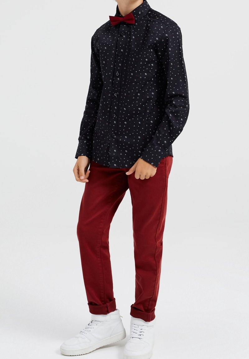 Camisa negra de botones con un patrón de puntos blancos, combinada con pantalones rojo oscuro y zapatillas blancas; incluye una pajarita roja como acento.
