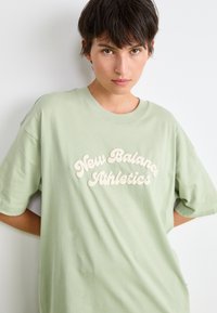 T-shirt en coton vert clair à coupe oversized et manches courtes. Présente un texte en relief blanc : « New Balance Athletics » sur la poitrine.