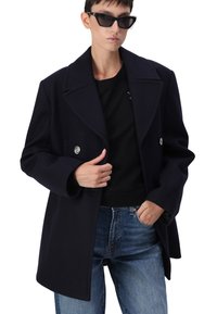 Jūras spēku zila vilnas maisījuma divrindu mētelis ar lielu apgriezto apkakli, sudraba krāsas pogām un brīvāku piegriezumu, apvienots ar melnu blūzi un džinsiem.