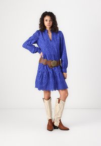 Blå klänning med ögla och långa ärmar, med en scalloped nederkant, parat med ett brunt läderbälte och krämfärgade cowboystövlar med bruna detaljer.