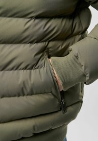 Ruka umetnuta u zatvoren džep maslinasto zelene podložene jakne s rebrastim rubom na rukavu.