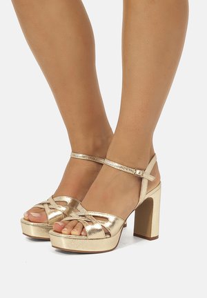 Par de sandalias de plataforma brillantes doradas con tacón alto y correas en el tobillo, usadas en los pies sobre un fondo blanco.