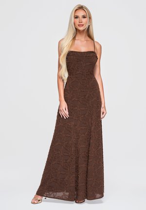 Femme aux longs cheveux blonds porte une robe maxi marron sans manches texturée et des talons ouverts dorés, debout devant un fond clair uni.