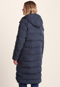 Woman with blonde hair wearing a long navy blue padded coat with a hood, viewed from the back, standing against a neutral background.