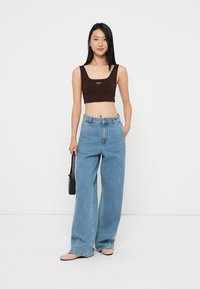 Brun cropped tank topp med logo, paret med høy midje lys blå vide bukser. Modellen holder en liten svart håndveske.