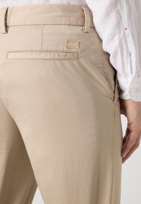 Bežīgas bikses, kas izgatavotas no gludas auduma, ar aizmugurējo kabatu ar izšūtu logotipa plombu un sānu kabatu. Veidotas fitā ar vienkāršu dizainu.