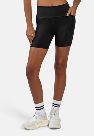 Frau trägt schwarze Biker-Shorts mit hohem Bund und Seitentasche, weiße Sportschuhe und weiße Socken mit schwarzen Streifen vor weißem Hintergrund.