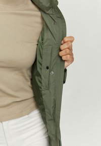 Olive green jacket with a smooth, textured exterior, featuring a side pocket and snap buttons, worn over a beige top.