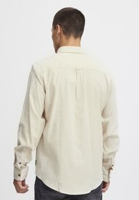 Chemise beige clair à manches longues avec un col à boutons, tissu texturé et détail de pli au dos. Manches à boutons avec des boutons contrastants.