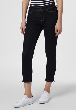 Mujer con vaqueros pitillo negros recortados, zapatillas blancas y una camiseta de rayas en blanco y negro, de pie frente a un fondo sencillo.