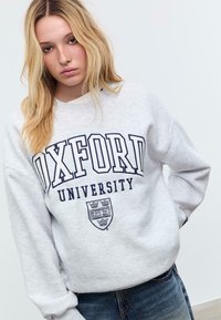 Mlada žena s dugom plavom kosom, obučena u svijetlu sivu duksericu Oxford University i plave traperice, naspram prazne bijele pozadine.