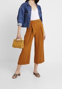 Kvinna i korta vida rostfärgade byxor, vit linne, blå denimjacka, sandaler med leopardmönster, och håller en liten gul vävd handväska.