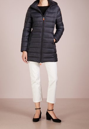 Kvinde iført en sort quiltejakke til midt på låret med lynlås, hvide korte bukser og sorte ankelrem-sandaler, stående på beige gulv.