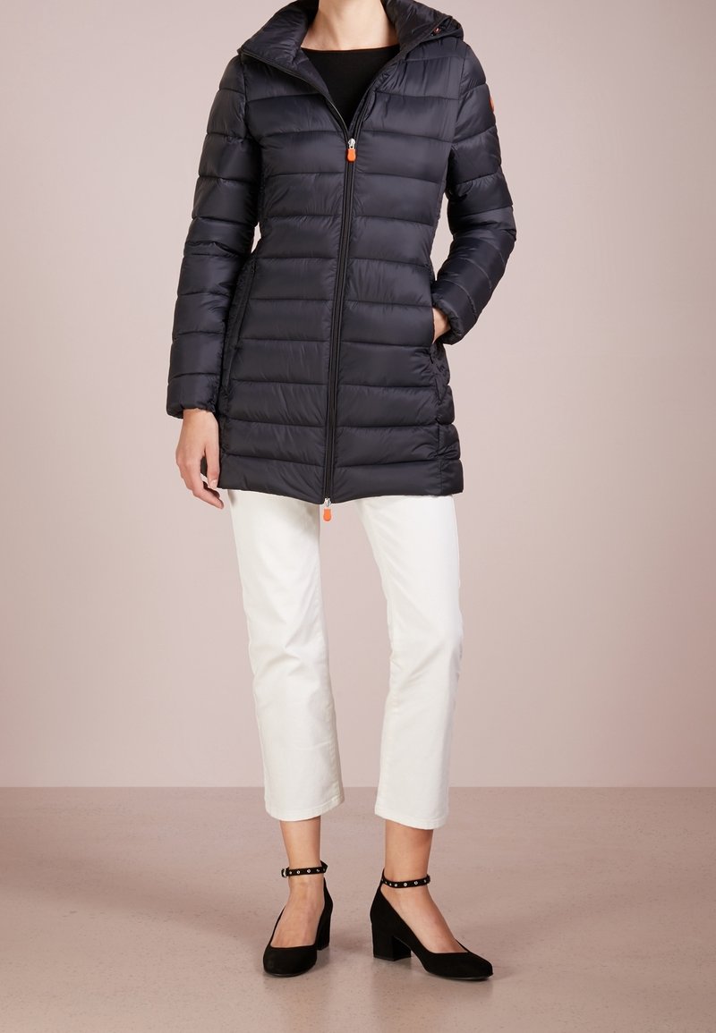 Femme portant une veste matelassée noire jusqu'à mi-cuisse avec fermeture éclair, un pantalon blanc court et des talons à bride cheville noirs, debout sur un sol beige.